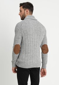Grå ribbestrikket genser med sjalkrage, med brune semskede albueputer. Laget av et teksturert strikkemateriale, tettsittende design.
