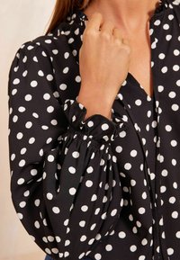 Blusa nera a pois bianchi, con colletto arricciato e polsini raccolti, realizzata in un tessuto leggero e textured.