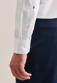 Biała koszula z długim rękawem i okrągłym mankietem z dwoma guzikami, z czarnym haftowanym wzorem róży na lewym rękawie. Widoczne granatowe spodnie.