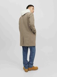 Beige Wollmantel mit einer grauen Kapuzenjacke, die hervorschaut, getragen über blauen Jeans und braunen Stiefeln. Der Mantel hat eine taillierte Passform und seitliche Taschen.