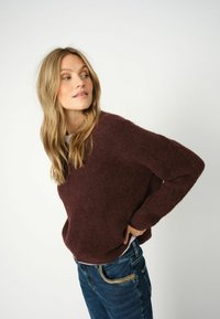 Bordeaux gebreid sweater met een relaxed fit en een diepe V-hals, gecombineerd met blauwe spijkerbroek. Zachte textuur en losse mouwen verhogen het comfort.