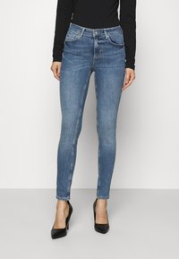 Jeans ajustados de mezclilla azul, con cintura de medio tiro, sutiles desgastes y diseño de cinco bolsillos, combinados con tacones negros puntiagudos.