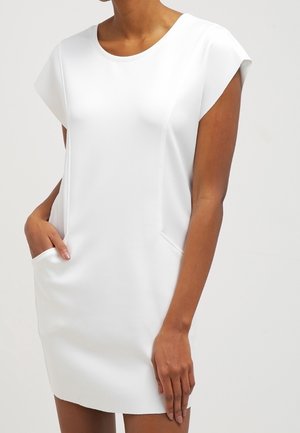 Mujer con un vestido blanco corto con mangas de capa y bolsillos, de pie con una mano en un bolsillo sobre un fondo liso.