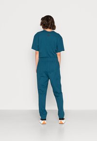 Persoon staat met zijn gezicht naar een witte muur, draagt een teal shirt met korte mouwen en bijpassende broek, en zwarte en witte sneakers.