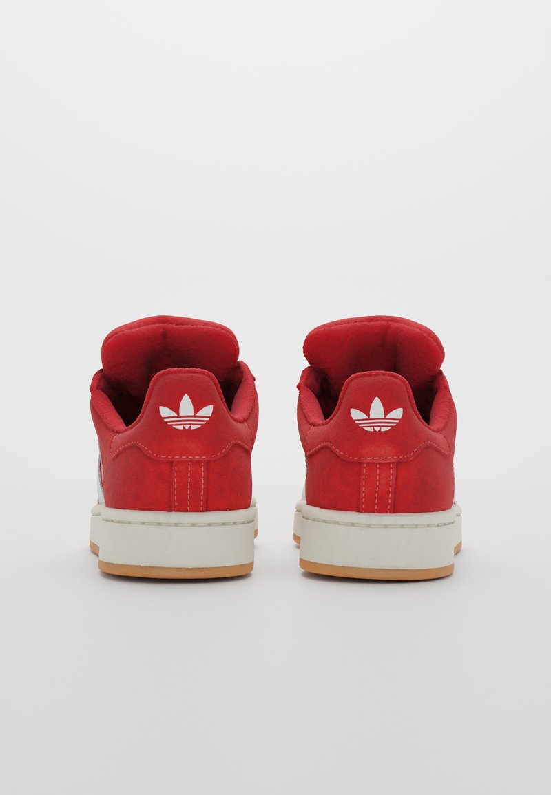 Czerwone zamszowe sneakersy z wyściełanymi kołnierzami, białym logo trefoil na piętach, białymi podeszwami środkowymi i gumowymi podeszwami zewnętrznymi. Klasyczny design sportowy.