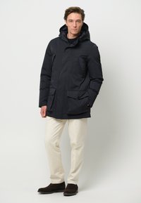 Marinblå isolerad parkas med huva, med två framfickor och dragkedja. I kombination med ljusa byxor och mörka stövlar.