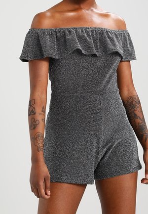 Femme portant une combinaison short grise texturée à épaules dénudées, manches courtes, avec détail volanté et tatouages visibles sur les deux avant-bras.