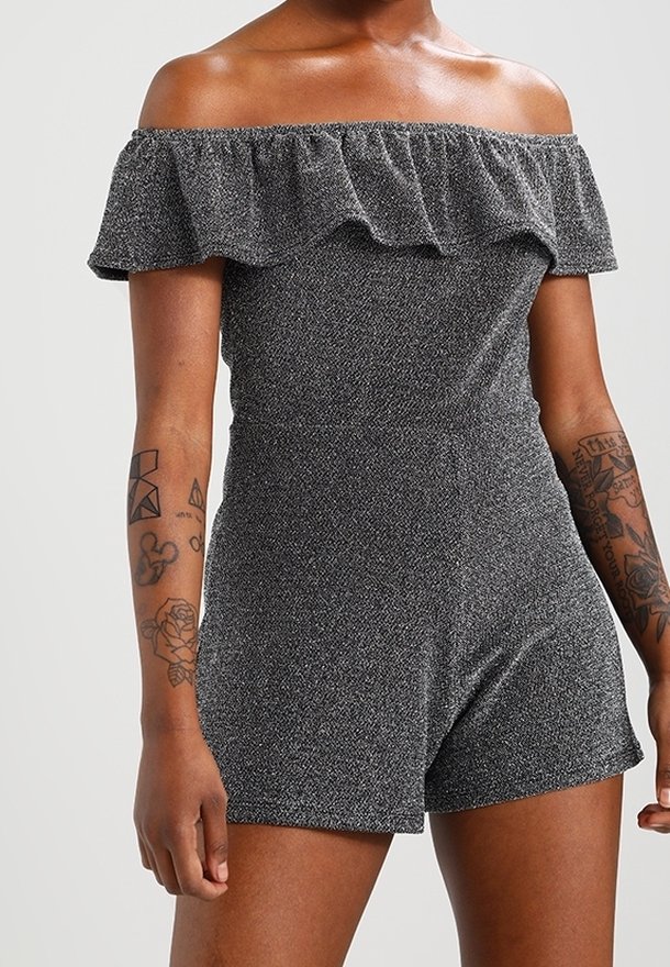 Femme portant une combinaison short grise texturée à épaules dénudées, manches courtes, avec détail volanté et tatouages visibles sur les deux avant-bras.