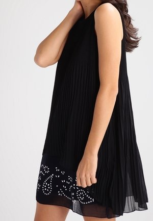 Frau in einem ärmellosen schwarzen Faltenkleid mit einem gemusterten, transparenten Saum, die sich vor einem schlichten hellen Hintergrund an den Hals fasst.