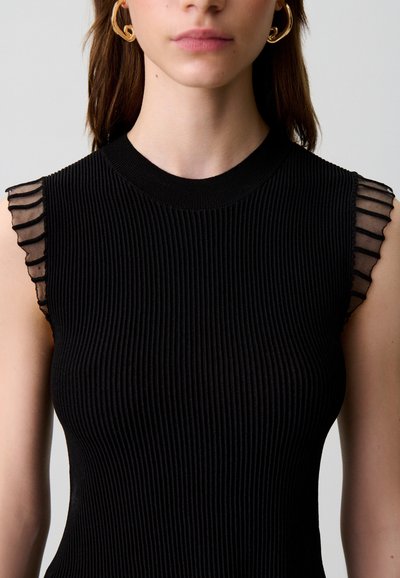 Donna che indossa un top nero a coste senza maniche con dettagli trasparenti arricciati sulle spalle e grandi orecchini a cerchio dorati, mostrata dal collo alla vita.