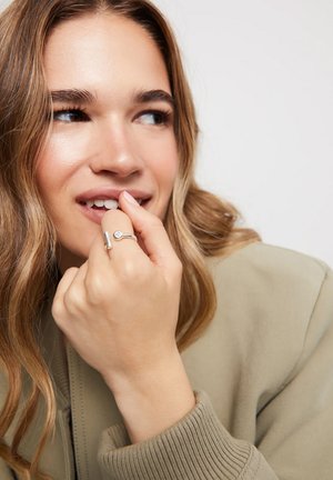Anillo de plata con una gema circular, llevado en una mano con puños de suéter beige. El diseño presenta una forma sencilla y moderna, y un acabado pulido.