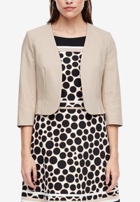 Femme portant un blazer beige court par-dessus une robe sans manches à pois noirs sur fond clair, debout devant un fond blanc.