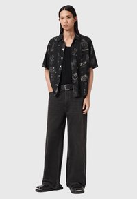 Μαύρο κοντομάνικο πουκάμισο με λευκά γραφικά μοτίβα, συνδυασμένο με φαρδιά μαύρα τζιν και χοντρά μαύρα παπούτσια. Αξεσουάρ περιλαμβάνουν δαχτυλίδι και ζώνη.