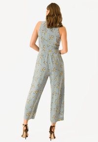 Blomstret jumpsuit i lyseblåt stof med tætsiddende overdel, vide ben og lynlås bagpå. Fremhævet med gule og hvide blomsterprint.