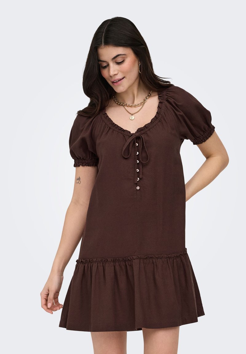 Femme portant une robe courte marron avec des manches bouffantes, des boutons sur le devant et un ourlet volanté, accessoirisée avec des colliers en or et des boucles d’oreilles créoles.