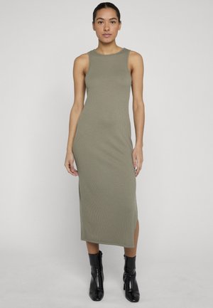 Femme debout, face à l'avant, portant une robe midi sans manches vert olive et des bottines noires, sur un fond clair uni.