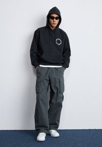 Hoodie noir avec poche avant et détail de logo, associé à un pantalon cargo gris avec poches latérales et des baskets blanches.
