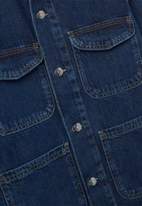 Giacca di denim blu scuro frontale con quattro tasche con alette e bottoni a pressione metallici disposti verticalmente al centro.