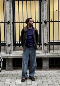 Homme portant une veste en peau lainée foncée et un jean ample se tient sur le trottoir devant une clôture en fer noire et un bâtiment jaune.