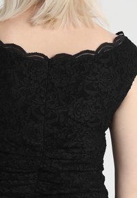 Svart spetsklänning med vågig halsringning; texturerat blommönster och rynkade detaljer framhäver form och design.