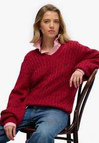 Jeune femme portant un pull en laine rouge à mailles torsadées et une chemise rose à col, assise sur une chaise en bois devant un fond blanc.