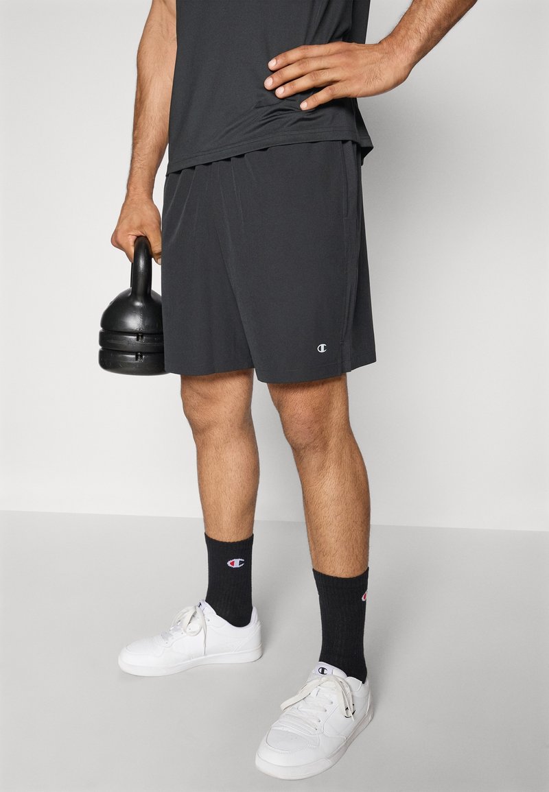 Czarne sportowe szorty z lekkiego materiału z bocznymi kieszeniami. Połączone z czarnymi skarpetkami i białymi sneakersami. Trzyma czarny odważnik kettlebell.