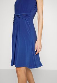 Kvinna iklädd en knälång klänning i kungligt blått med en tvinnad knutdetalj i midjan, stående mot en enkel ljus bakgrund.