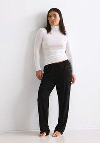 White long-sleeve turtleneck top paired with loose black pants, both made from soft, stretchy fabric. Barefoot on a light surface.