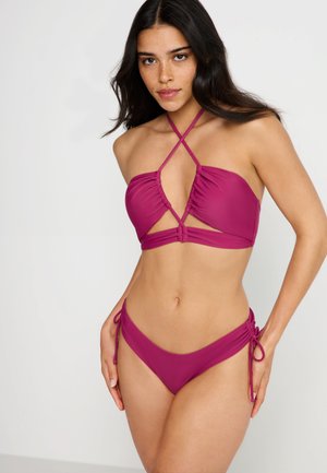 Magenta-Bikini mit Halteroberteil mit Kreuzträgern, das über geraffte Details und Ausschnitte verfügt, kombiniert mit einem verstellbaren, seitlich zu bindenden Höschen. Glatte Textur.