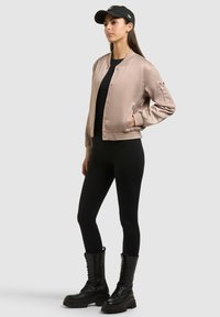 Femme portant une veste bomber en satin beige, des leggings noirs, des bottes noires à lacets et une casquette de baseball noire, debout avec une main dans la poche.