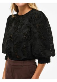 Svart teksturert topp med puffede ermer, med et lagdelt mønster og myke, frynsete kanter. Kombinert med høyvidde brune bukser.