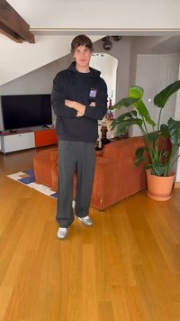 Černá mikina s fialovým logem, šedé kalhoty širokého střihu a stříbrné boty. Vnitřní prostředí s rostlinou a pohovkou s oranžovými pruhy.