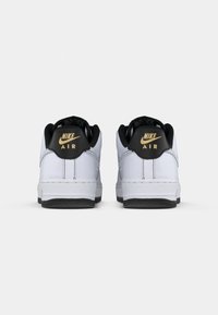 Et par hvite Nike Air-sneakers med svarte hællapper og gull Nike Air-logoer, sett fra bak på en enkel hvit bakgrunn.