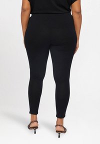 Czarne legginsy wykonane z elastycznego materiału, z wysokim stanem i gładką fakturą. Noszone z leopardowymi szpilkami, ukazując widok z tyłu.