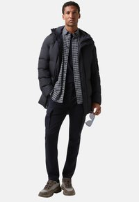 Puffjakke i mørk navy, lagdelt over en sort og hvid ternet skjorte. Sorte bukser med cargo-lommer og grå-brune sneakers.