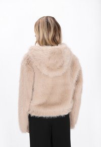 Korte nep-bont jas in lichtbeige, met een capuchon en lange mouwen. De textuur is zacht met een pluizige uitstraling, gecombineerd met zwarte onderkleding.