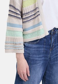 Strickjacke mit mehrfarbigen Streifen in Creme, Blau, Grün und Marineblau, mit strukturierten Mustern und Glockenärmel, kombiniert mit blauen Jeans.