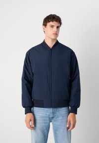 Veste de bombardier navy avec une fermeture éclair à l'avant, des poignets et une taille côtelés. Matériau lisse et coupe décontractée, associée à un jean bleu clair.