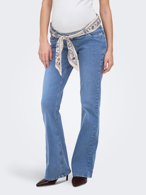 Kvinde iført lyseblå bootcut jeans med cremefarvet mønstret tørklæde som bælte og mørke spidse høje hæle, stående mod hvid baggrund.