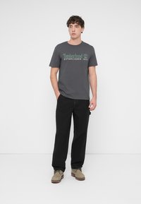 Grijs katoenen t-shirt met groene tekst "Timberland Established 1973", gecombineerd met een zwarte broek en lichtbruine suède schoenen.