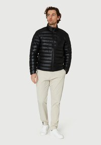 Sort puffjakke med lynlås foran, horisontal quiltet design, høj krave og en brystlomme, parret med beige bukser og hvide sneakers.