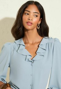 Femme avec de longs cheveux sombres portant des boucles d'oreilles en or et une blouse bleu clair avec un col volanté et des boutons, regardant sur le côté.