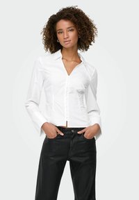 Chemise blanche ajustée avec col, manches longues et fermeture à boutons sur le devant. Associée à un jean noir, créant une tenue contrastante.