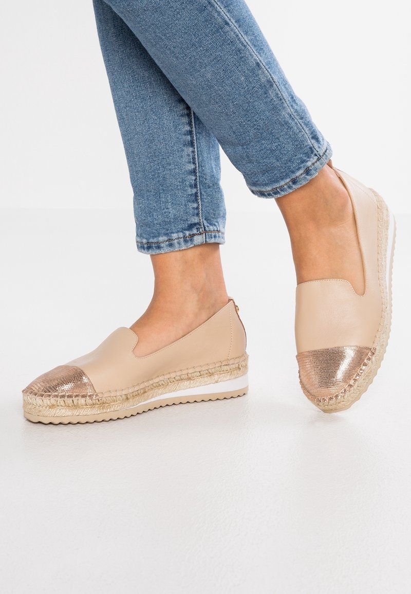 Espadrilles beige à enfiler avec des embouts en rosegold métallique, portées avec un jean bleu retroussé sur un sol blanc.