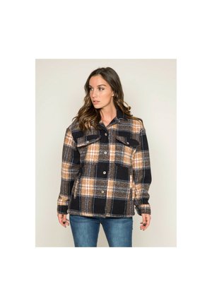 Femme portant une veste à carreaux noire, beige et orange avec des boutons, par-dessus un jean bleu, regardant vers sa gauche sur un fond uni.