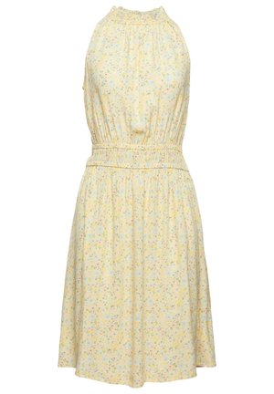 Personne portant une veste matelassée beige par-dessus une robe jaune fleurie à longueur de genou avec des sandales à lanières tressées beige, debout devant un fond uni.