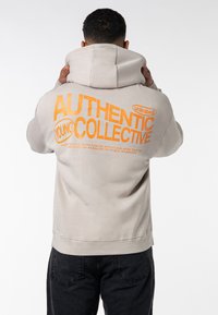 Beige hoodie with a large orange graphic text on the back reading "AUTHENTIC YOUNG COLLECTIVE." Features a drawstring hood and kangaroo pocket.