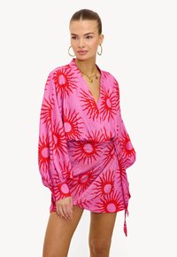 Vestido cruzado rosa con patrón de estallido solar rojo, mangas largas y un escote en V profundo. Cuenta con un lazo lateral y tela ligera. Accesorios de joyería dorada.