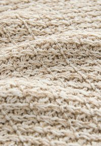 Tissu tricoté beige présentant un motif texturé avec des points en relief, créant une apparence tissée rythmique. Surface douce et texturée avec une variation tonale subtile.