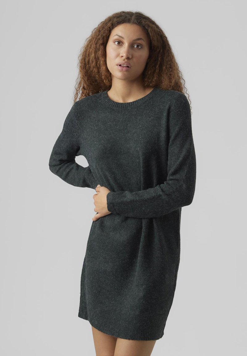 Junge Frau mit lockigem Haar trägt ein dunkelgraues langärmliges Strickkleid und steht mit einer Hand in der Hüfte und der anderen an der Taille.